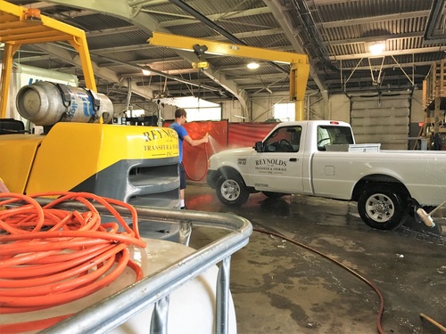 A person washes a white Reynolds company pickup truck inside a large garage. A yellow forklift, red hose, and other cleaning equipment are visible in the foreground. The area is well-lit with open space and industrial fixtures.