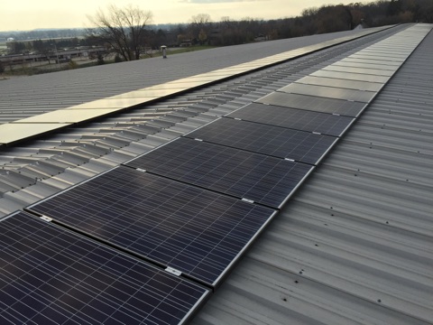 Rows of solar panels are installed on a metal rooftop, stretching into the distance. Trees and a river are visible in the background under a partly cloudy sky.